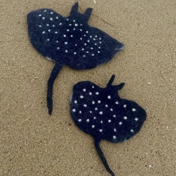 Two dark-blue wool felted stingray shapes with white spots lying on a sandy beach.