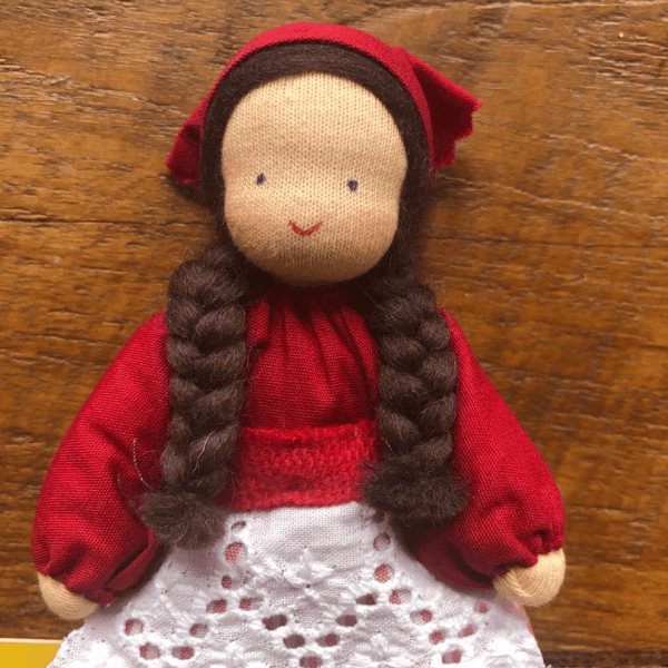 IMG_1832 | Bambino Eco Toys Soft cloth doll with long braided brown hair, wearing a red dress, red headscarf, and white lace apron, smiling against a wooden background.