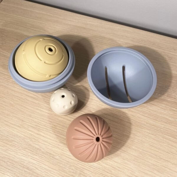 Set of pastel silicone parts on a wooden surface: two hollow blue hemispheres, a beige dome with a central hole, a white dotted bead, and a pink ridged sphere; components appear to assemble into a spherical toy or organizer.