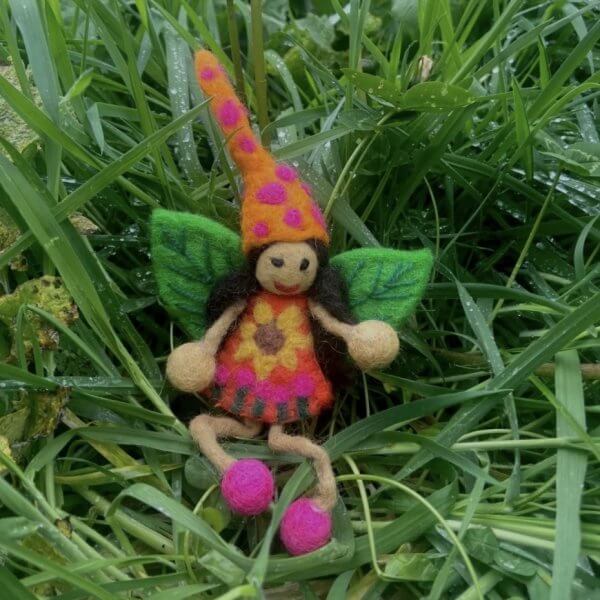 Screenshot | Bambino Eco Toys Colorful felt fairy toy with orange hat, pink spots, yellow floral dress, green felt wings, and pink shoes perched in green grass.
