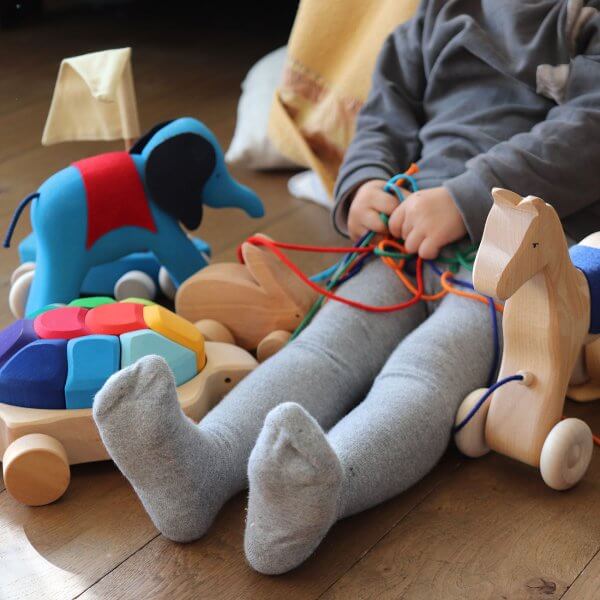 Child sitting on a wooden floor playing with colorful string toys and wooden animal toys, wearing gray leggings and socks.